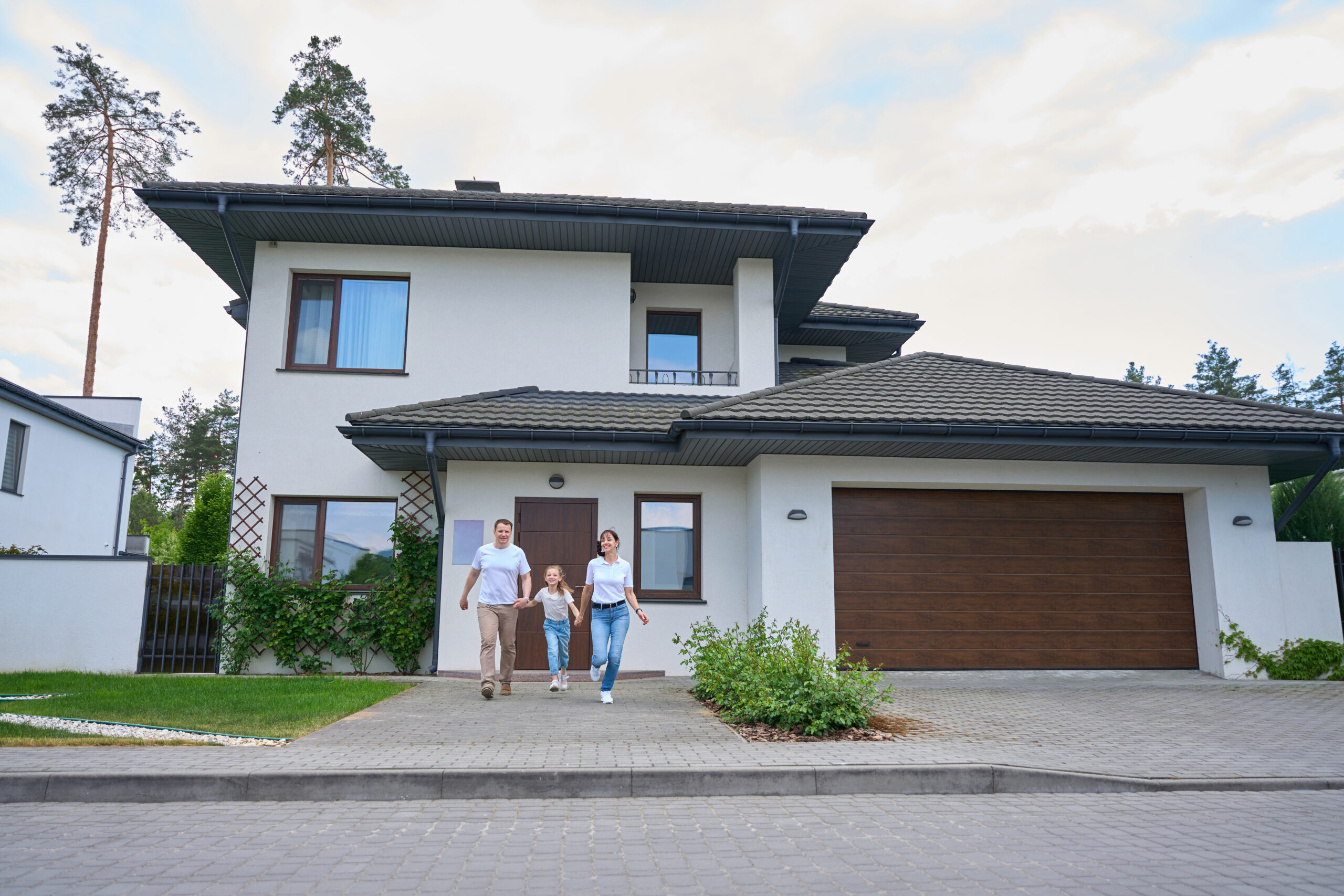 Happy caucasian mother, father and daughter holding hands and running from new modern townhouse outdoors in cloudy day. Family future planning, relationship and enjoying time together